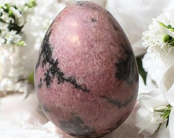 May include: A polished, egg-shaped stone with a pink and black marbled pattern. The stone is smooth and reflective, with a natural, organic appearance. The background features white flowers and soft, white fabric.