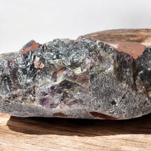 May include: A large, rough, dark grey and purple amethyst crystal with a reddish-brown hue. The crystal has a natural, irregular shape and is resting on a wooden surface.