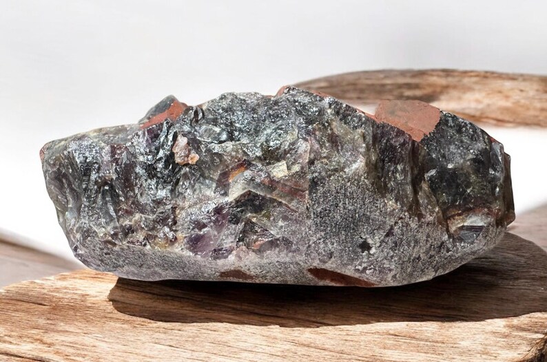 May include: A large, rough, dark grey and purple amethyst crystal with a reddish-brown hue. The crystal has a natural, irregular shape and is resting on a wooden surface.