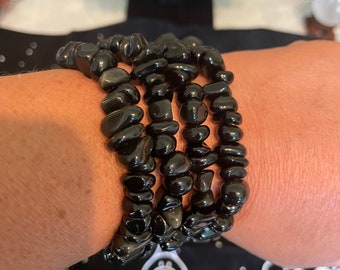May include: Four black gemstone bracelets stacked on a person's wrist. The bracelets are made of small, irregular shaped black stones.