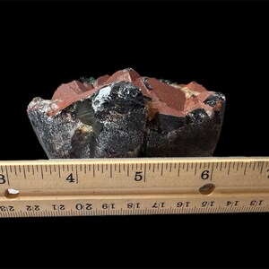 K&ouml;nnte beinhalten: Nahaufnahme eines dunkelroten, schwarzen und gr&uuml;nen Kristallclusters. Der Kristall ist etwa 12,7 cm breit und liegt auf einem Holzlineal.