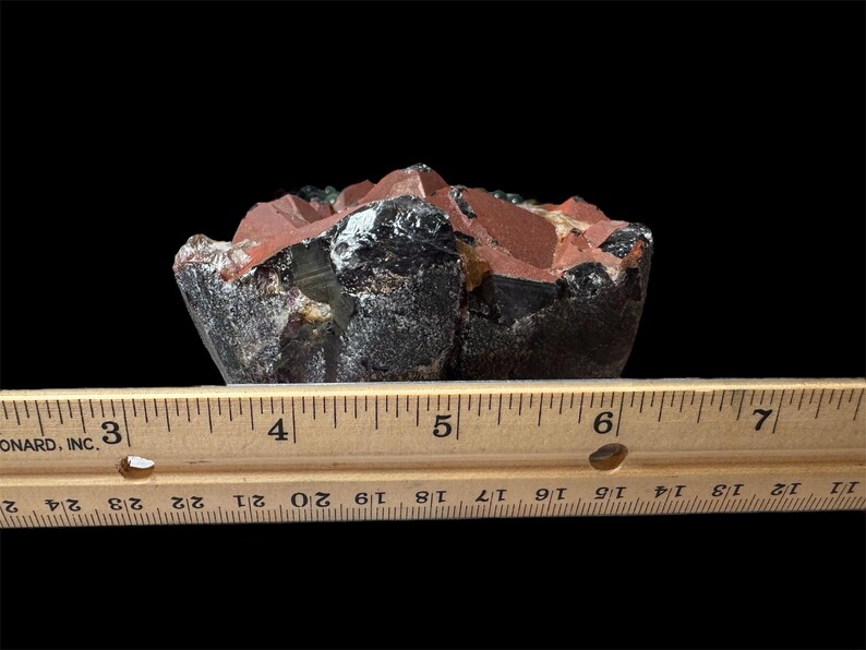 K&ouml;nnte beinhalten: Nahaufnahme eines dunkelroten, schwarzen und gr&uuml;nen Kristallclusters. Der Kristall ist etwa 12,7 cm breit und liegt auf einem Holzlineal.