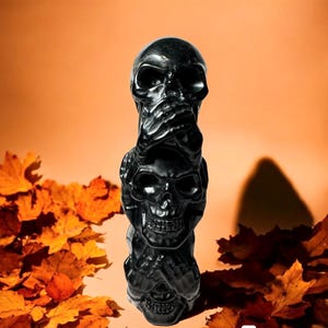 May include: A black sculpture of three stacked skulls, each with its hands covering its eyes, mouth, and ears. The sculpture is set against a backdrop of orange and brown fall leaves.