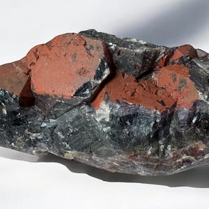 May include: A large, dark gray and purple amethyst crystal with red inclusions. The crystal has a rough, natural texture.