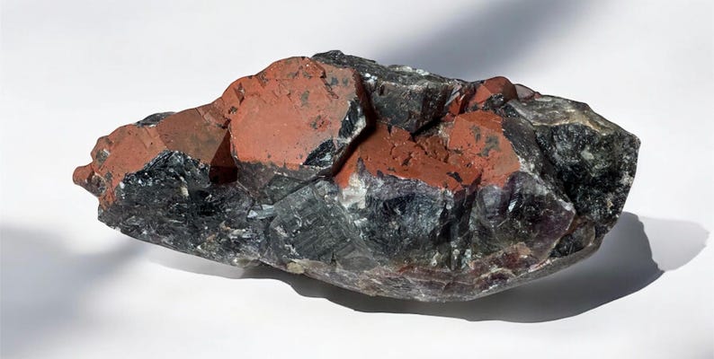 May include: A large, dark gray and purple amethyst crystal with red inclusions. The crystal has a rough, natural texture.