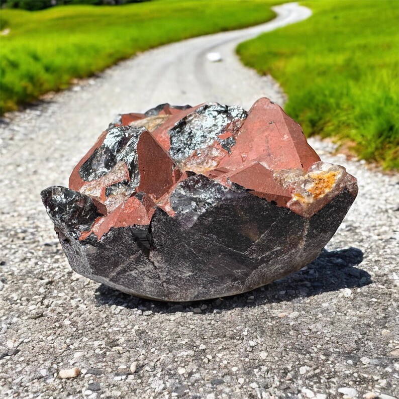 K&ouml;nnte beinhalten: Ein gro&szlig;er, dunkelroter und schwarzer Kristallcluster mit einer rauen, nat&uuml;rlichen Textur. Der Kristall liegt auf einem Schotterweg.