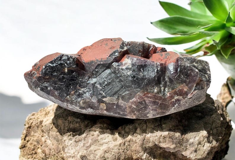 May include: A large, dark purple amethyst crystal cluster with red inclusions. The crystal is rough and has a natural, raw appearance. It is sitting on a brown rock.