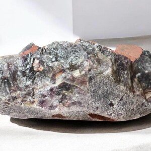 May include: A large, irregularly shaped amethyst crystal with a purple and grey color. The crystal has a rough, natural texture and is partially translucent.