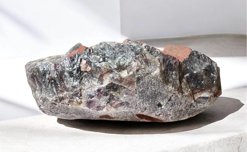 May include: A large, irregularly shaped amethyst crystal with a purple and grey color. The crystal has a rough, natural texture and is partially translucent.