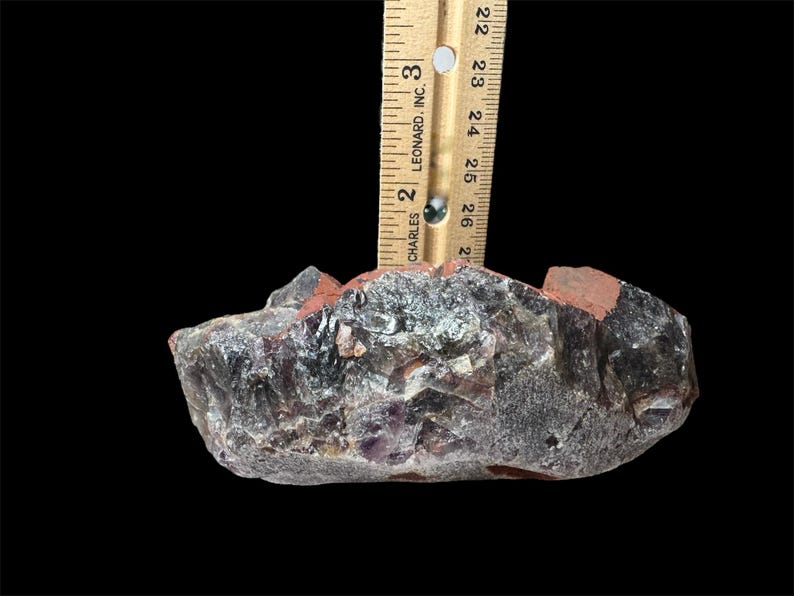 May include: A large, rough amethyst crystal with a purple and gray color. The crystal is approximately 2.5 inches wide and has a natural, irregular shape.