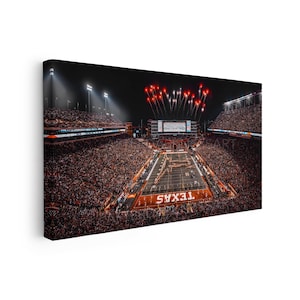 May include: A panoramic canvas print of a night football stadium filled with spectators. The field features the word TEXAS in the end zone, bright stadium lights, and red fireworks exploding in the dark sky above the scoreboard.