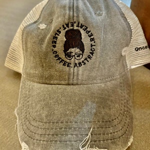 May include: A gray distressed trucker hat with a black embroidered design of a woman with a bun, glasses, and the text "Coffee. Abstract.  Live. Repeat. 42375".