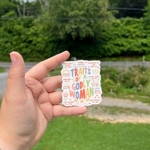 May include: A colorful sticker with the words "Traits of a Godly Woman" in various fonts and colors. The sticker includes words like "confident," "patient," and "trustworthy," with associated Bible verses. The background is a mix of white and pastel colors.