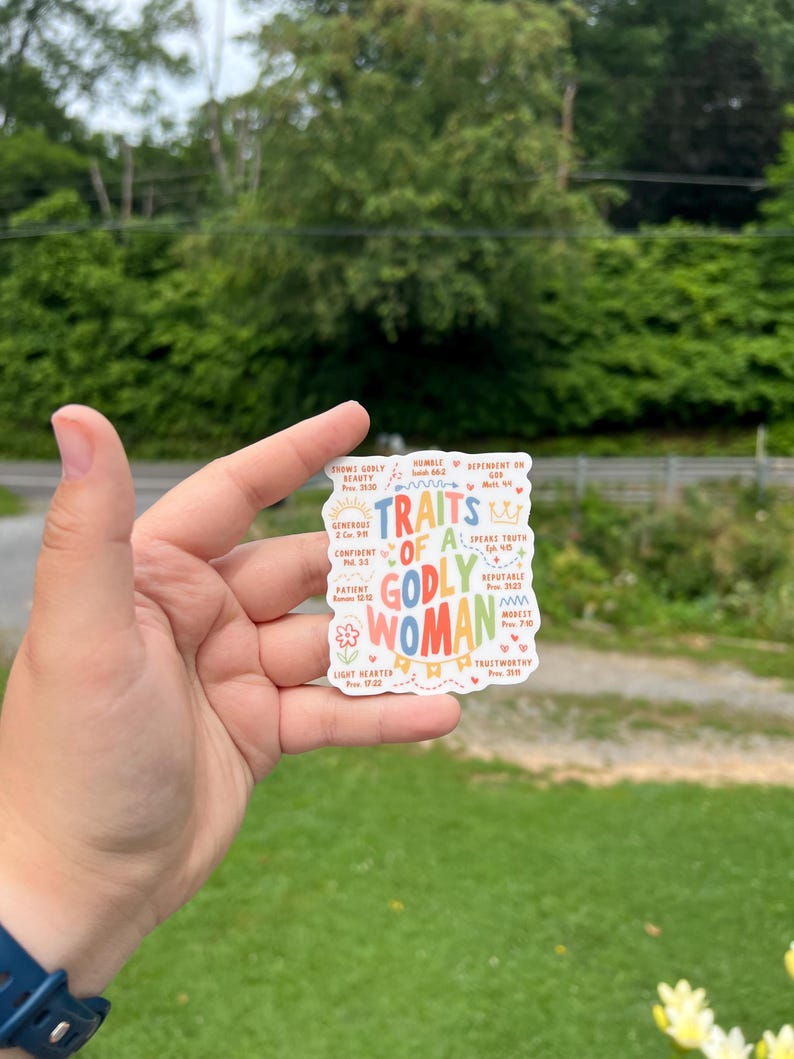 May include: A colorful sticker with the words "Traits of a Godly Woman" in various fonts and colors. The sticker includes words like "confident," "patient," and "trustworthy," with associated Bible verses. The background is a mix of white and pastel colors.