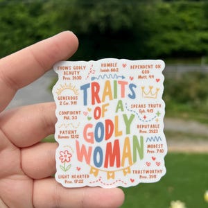 May include: A white sticker with the words "TRAITS OF A GODLY WOMAN" in colorful letters. The sticker lists various traits such as "Shows Godly Beauty," "Humble," and "Trustworthy," with corresponding Bible verses. The sticker is held by a hand.