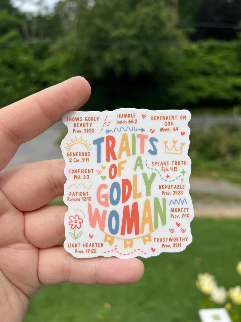 May include: A white sticker with the words "TRAITS OF A GODLY WOMAN" in colorful letters. The sticker lists various traits such as "Shows Godly Beauty," "Humble," and "Trustworthy," with corresponding Bible verses. The sticker is held by a hand.