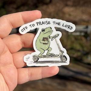 May include: A sticker featuring a green frog riding a scooter and holding a book. The text "OFF TO PRAISE THE LORD" arches above the frog. The sticker is held in a hand against a blurred outdoor background.