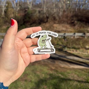 May include: A white sticker featuring a cartoon frog riding a scooter and holding a book. The frog is green, and the scooter is black and white. The text "OFF TO PRAISE THE LORD" is above the frog.
