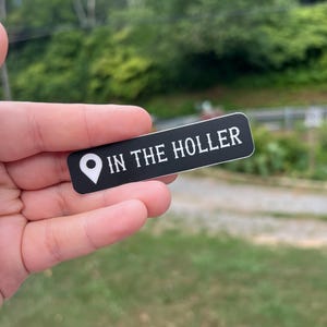 Pode incluir: Um adesivo preto com texto branco que diz "IN THE HOLLER" e um &iacute;cone de alfinete branco.