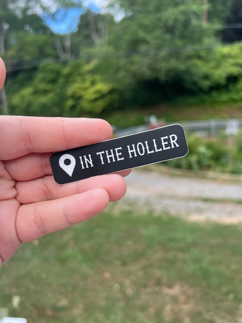 Pode incluir: Um adesivo preto com texto branco que diz "IN THE HOLLER" e um &iacute;cone de alfinete branco.