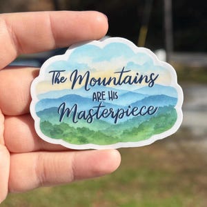 May include: A sticker with a cloud-shaped border featuring a watercolor mountain scene in shades of blue, green, and yellow. The text "The Mountains are His Masterpiece" is written in dark blue script.
