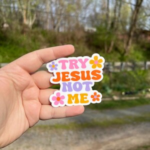 May include: A sticker with the text "Try Jesus Not Me" in a retro font with pink, orange, and purple colors. The sticker is surrounded by small pink, orange, and yellow flowers.