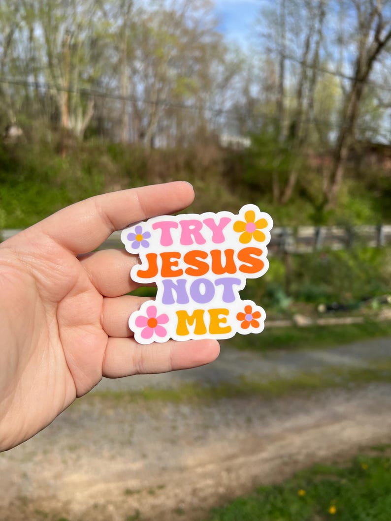 May include: A sticker with the text "Try Jesus Not Me" in a retro font with pink, orange, and purple colors. The sticker is surrounded by small pink, orange, and yellow flowers.