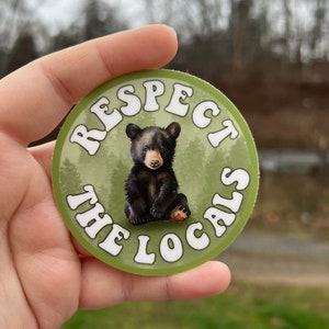 May include: A round sticker with a green background and white text that reads "Respect the Locals". A black bear cub is sitting in the centre of the sticker.