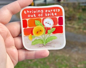 May include: A square sticker with the text "thriving purely out of spite" in white letters on a red and orange background. The sticker features a cartoon dandelion with a smiling yellow flower and a fluffy white seed head.