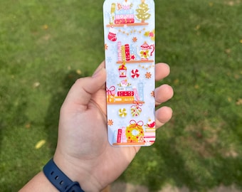 May include: A rectangular bookmark featuring a festive Christmas bookshelf design. The bookmark showcases shelves with books, a Christmas tree, and holiday decorations in red, white, and gold. The word "MERRY" is visible.