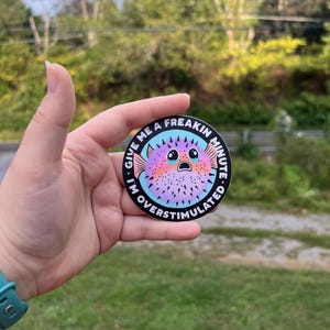 May include: A round sticker with a black background and white text that reads "Give me a freakin minute I'm overstimulated".  A cartoon pufferfish with pink, purple, and blue colors is in the center of the sticker. The pufferfish has big eyes and a surprised expression.