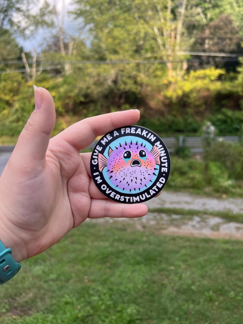 May include: A round sticker with a black background and white text that reads "Give me a freakin minute I'm overstimulated".  A cartoon pufferfish with pink, purple, and blue colors is in the center of the sticker. The pufferfish has big eyes and a surprised expression.