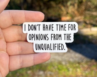 May include: A white sticker with black text that reads "I DON'T HAVE TIME FOR OPINIONS FROM THE UNQUALIFIED." The sticker is held in a hand with red nail polish, with a blurred green and brown background.