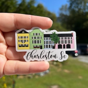 May include: A sticker featuring a row of colorful houses with a palm tree in front. The houses are yellow, green, and pink. The sticker also includes the text "Charleston, SC".