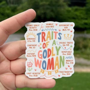 May include: A sticker with the words "Traits of a Godly Woman" in colorful letters. The sticker includes phrases like "Shows Godly Beauty," "Humble," and "Dependent on God," with corresponding Bible verses. The sticker is held by a hand.