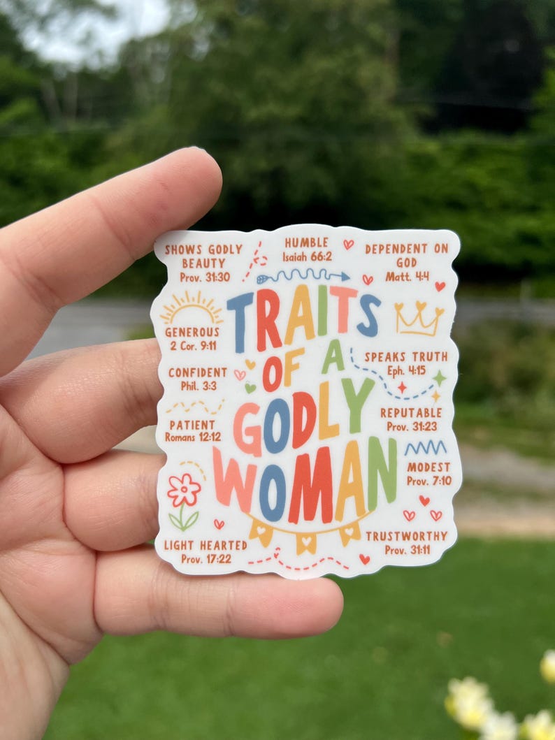 May include: A sticker with the words "Traits of a Godly Woman" in colorful letters. The sticker includes phrases like "Shows Godly Beauty," "Humble," and "Dependent on God," with corresponding Bible verses. The sticker is held by a hand.