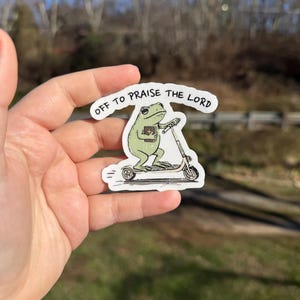May include: A sticker featuring a green frog riding a scooter and holding a book. The sticker has the text "OFF TO PRAISE THE LORD" in a white banner. The frog is light green with black eyes and is riding a white and gray scooter.