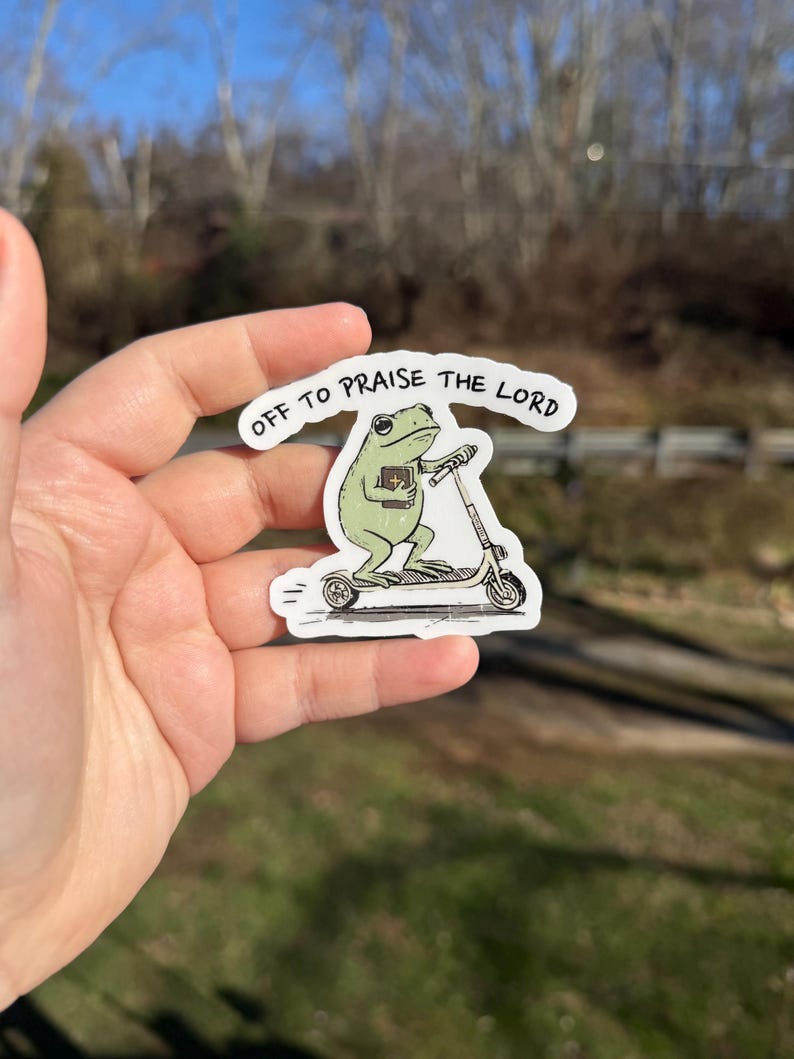 May include: A sticker featuring a green frog riding a scooter and holding a book. The sticker has the text "OFF TO PRAISE THE LORD" in a white banner. The frog is light green with black eyes and is riding a white and gray scooter.