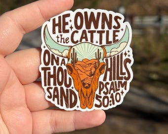May include: A sticker with the text "HE OWNS the CATTLE ONA THOUSAND HILLS PSALM 50:10" in a stylized font. The sticker features a brown steer skull with turquoise horns and a desert landscape design.