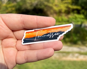 May include: A sticker of the state of Tennessee with a sunset over mountains in the background. The text "Tennessee" is written in white cursive across the sticker.