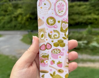 May include: A rectangular phone case featuring a repeating pattern of matcha-themed illustrations in pink, white, and gold. The design includes matcha lattes, strawberries, and tea-making tools.