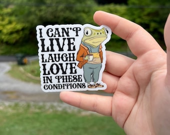 May include: A sticker featuring a cartoon frog wearing a brown jacket and holding a cup of coffee. The text on the sticker reads "I can't live laugh love in these conditions."