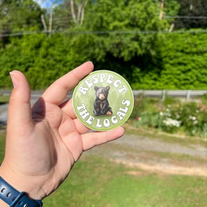 May include: A round sticker with a green background and white text that reads "Respect the Locals". A small black bear cub is sitting in the centre of the sticker.