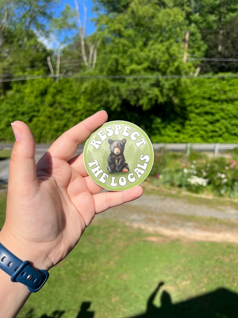 May include: A round sticker with a green background and white text that reads "Respect the Locals". A small black bear cub is sitting in the centre of the sticker.