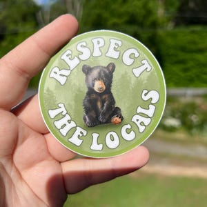 May include: A round sticker with a green background and white text that reads "Respect the Locals". A black bear cub is in the centre of the sticker.