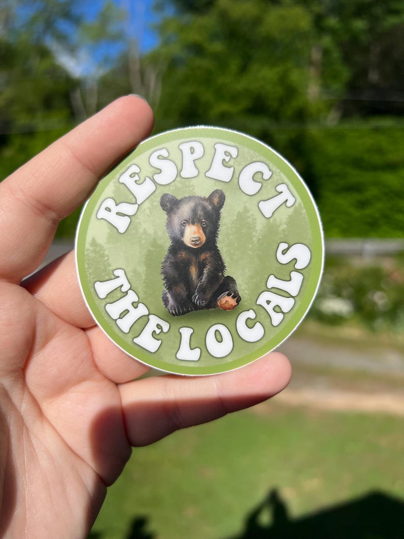 May include: A round sticker with a green background and white text that reads "Respect the Locals". A black bear cub is in the centre of the sticker.