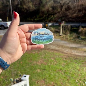 May include: A sticker with a cloud-shaped border featuring a mountain landscape in shades of blue and green. The text "The Mountains are His Masterpiece" is written across the top and bottom of the design.