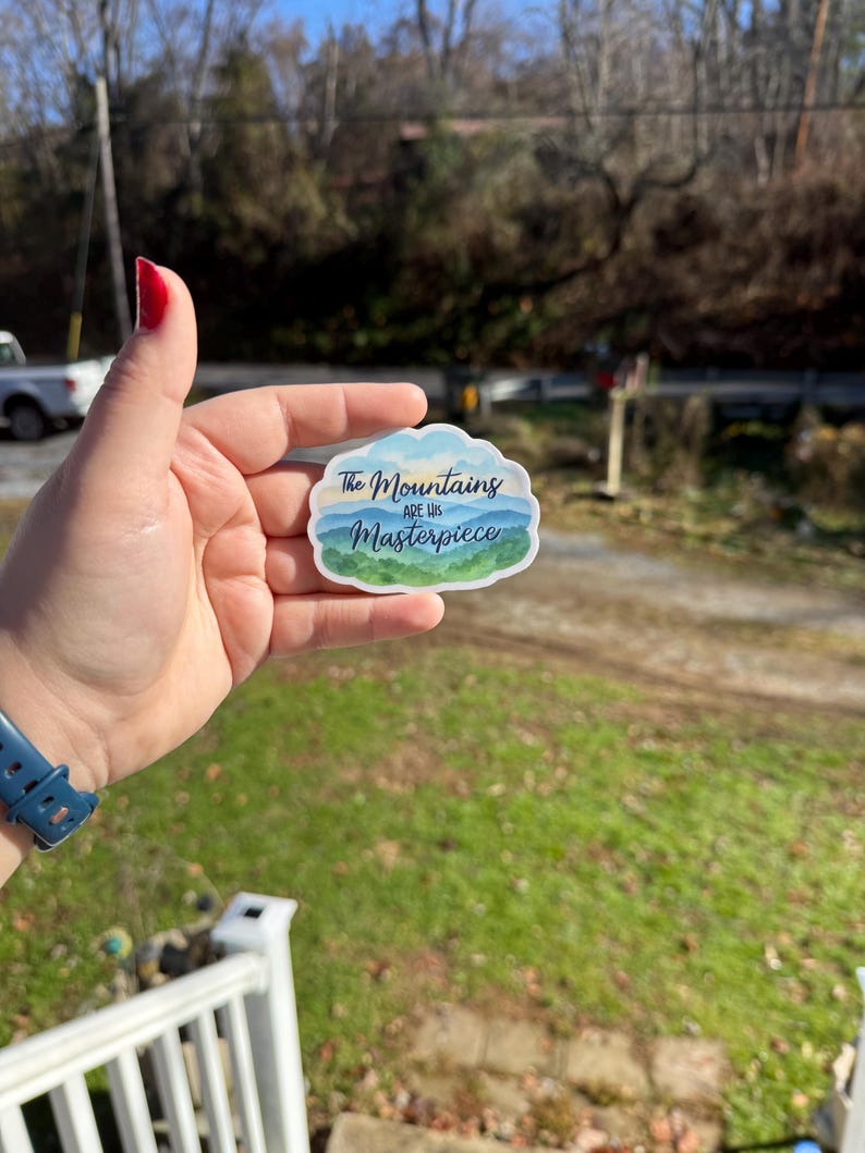 May include: A sticker with a cloud-shaped border featuring a mountain landscape in shades of blue and green. The text "The Mountains are His Masterpiece" is written across the top and bottom of the design.