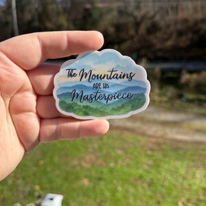 May include: A sticker with a cloud-shaped border featuring a mountain landscape design. The sticker has a blue, green, and white color scheme with the text "The Mountains are His Masterpiece" in a handwritten font.