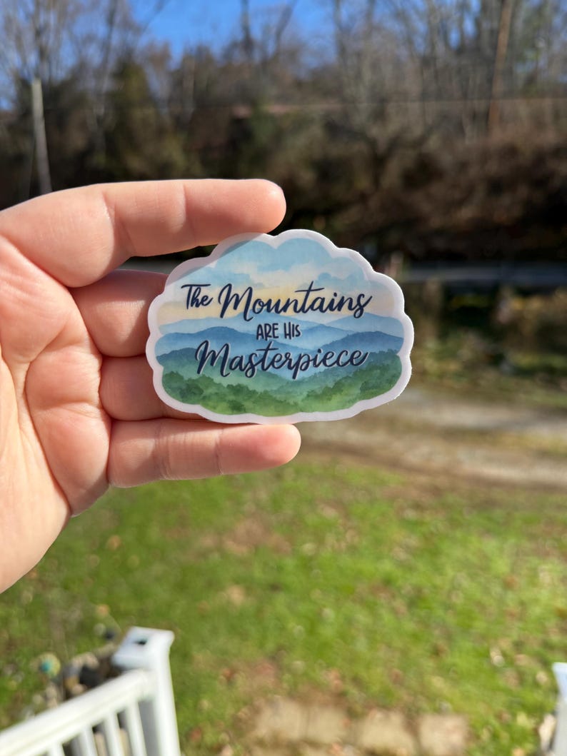 May include: A sticker with a cloud-shaped border featuring a mountain landscape design. The sticker has a blue, green, and white color scheme with the text "The Mountains are His Masterpiece" in a handwritten font.